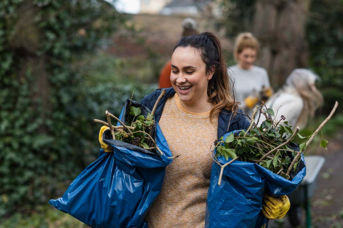 A front view of a young brunette woman carrying two bags of garden waste that is to be composted and reused. She is working on a community space garden in Hexham in the North East of England with a group of friends.